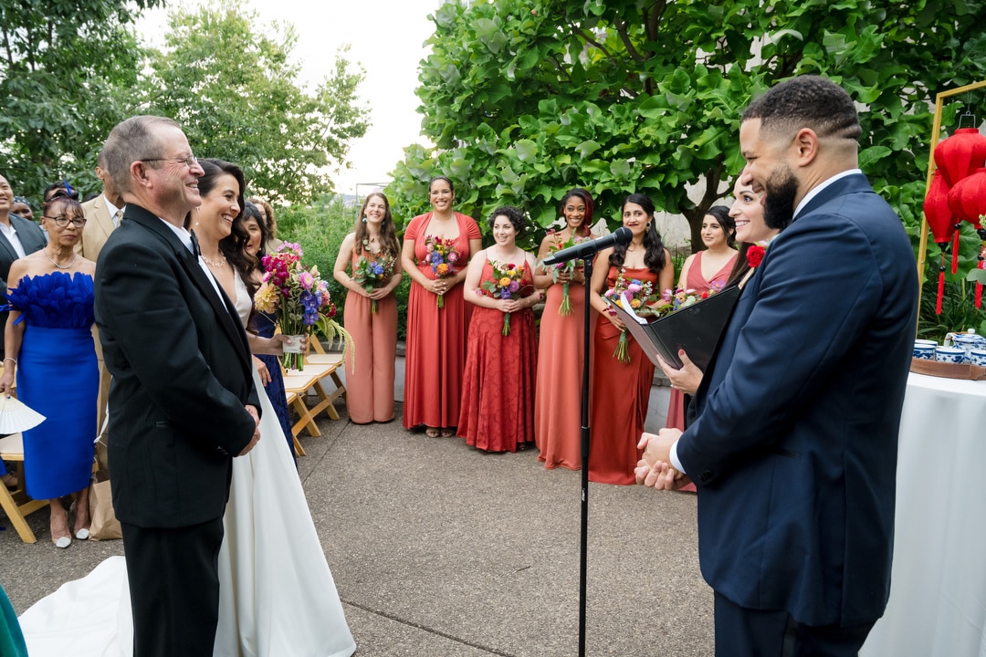 A bride and groom stand at the altar with an officiant during an outdoor wedding ceremony, surrounded by bridesmaids in matching dresses holding bouquets. Guests watch the smiling couple.