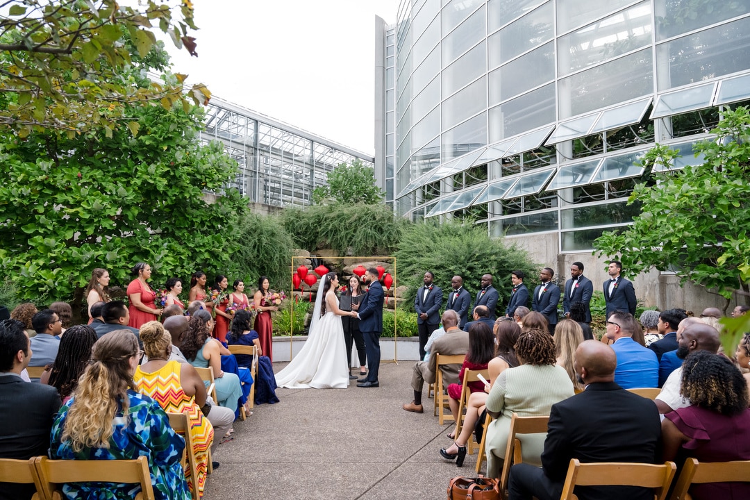 A bride and groom stand at the altar holding hands during an outdoor wedding ceremony, surrounded by bridesmaids, groomsmen, and seated guests, with a modern glass building and lush greenery as the backdrop.