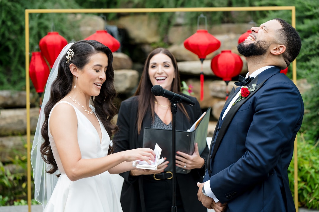 A bride and groom stand at an outdoor altar, laughing joyfully during their wedding ceremony. The officiant smiles behind them, and red lanterns hang in the background amid greenery.