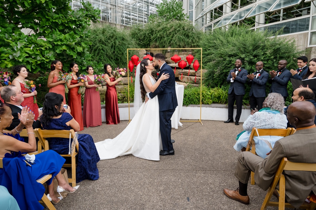 A bride and groom share their first kiss at an outdoor wedding ceremony, surrounded by applauding guests and a wedding party dressed in coordinated colors, with greenery and red lanterns in the background.