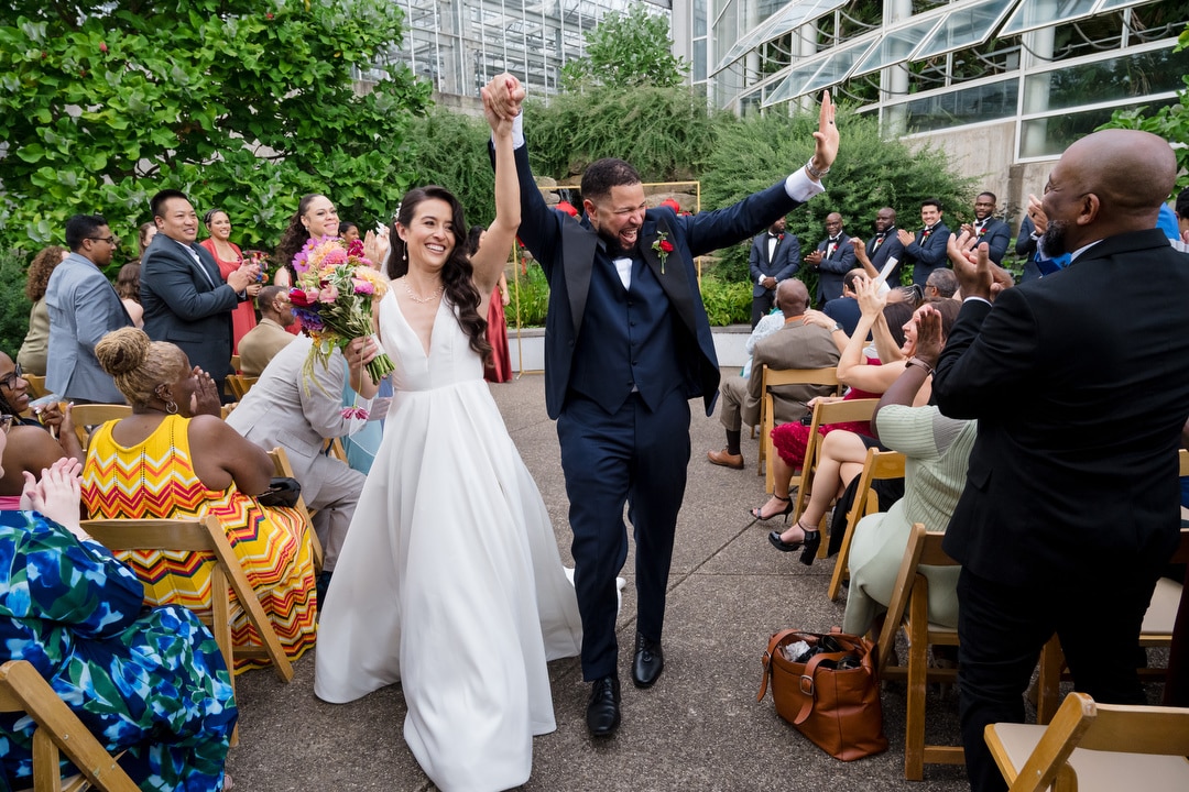 A bride and groom, smiling and holding hands raised in celebration, walk down the aisle outdoors as guests seated on both sides applaud and cheer. The bride holds a bouquet and wears a white dress; the groom is in a dark suit.
