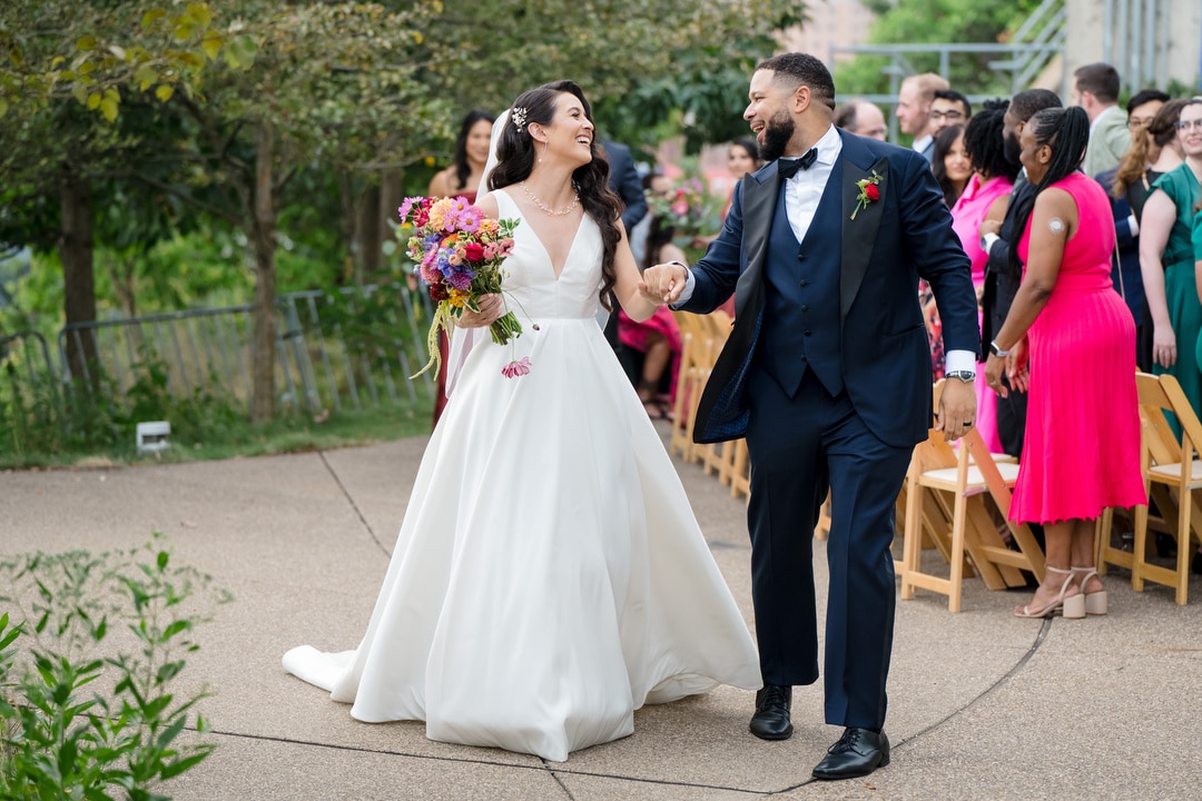 A bride in a white gown holding a colorful bouquet walks hand-in-hand with a groom in a navy tuxedo down an outdoor aisle, smiling at each other, as guests in bright attire look on and celebrate.