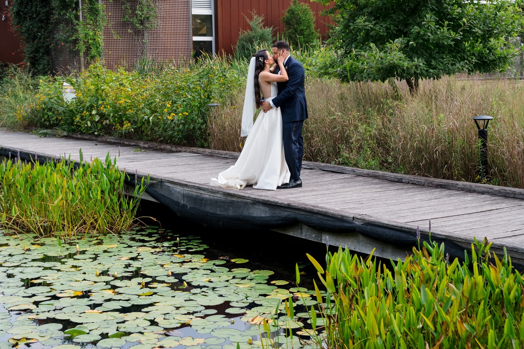 A bride and groom stand on a wooden boardwalk, surrounded by tall grass, trees, and water lilies, sharing an intimate moment outdoors on their wedding day.