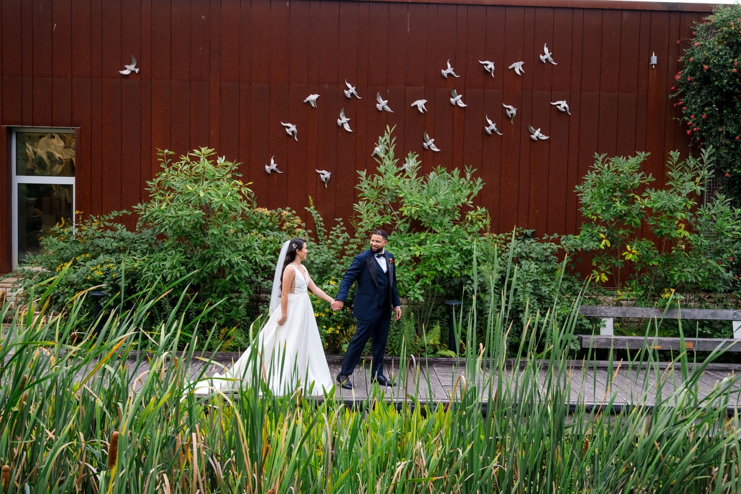 A bride and groom walk hand-in-hand on a wooden path surrounded by lush greenery. Behind them is a rust-colored wall with white bird sculptures in flight and a window on the left side.