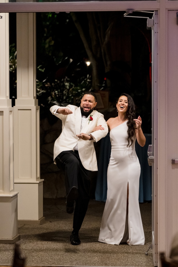 A joyful bride and groom in wedding attire excitedly enter a venue, smiling and laughing. The groom playfully raises one leg, while the bride holds his arm and beams.
