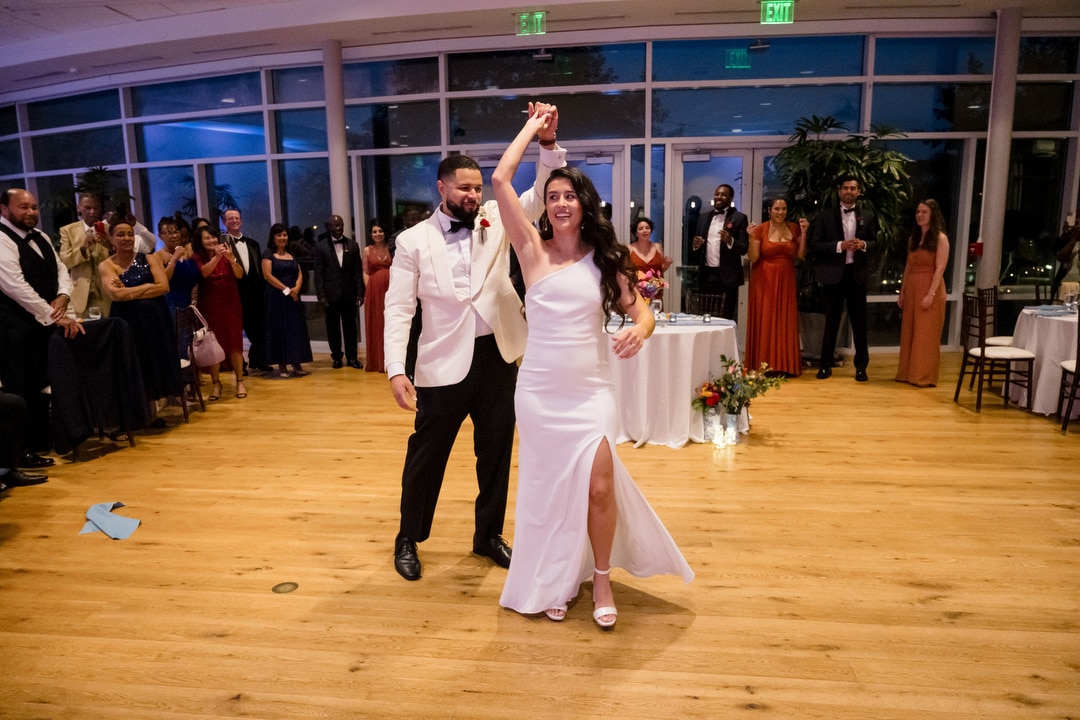 A bride and groom, both smiling, dance together on a wooden floor in a bright, modern venue, surrounded by guests watching and celebrating. The bride wears a white dress; the groom wears a white jacket and black pants.