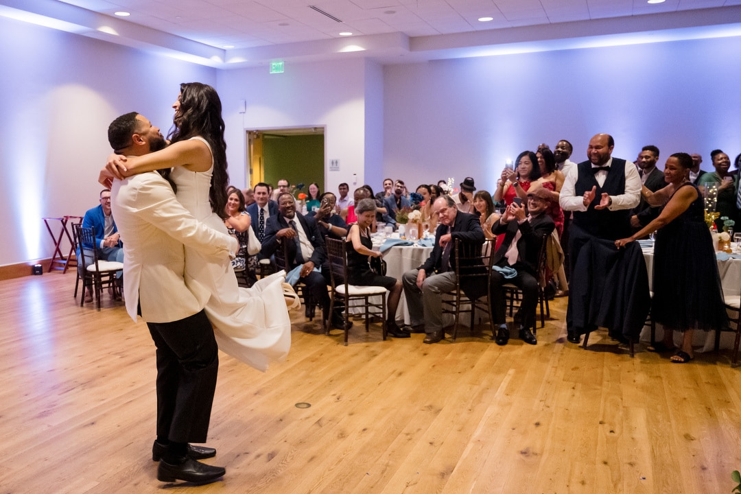 A groom lifts his bride joyfully on a dance floor while wedding guests seated at round tables watch and smile in a warmly lit reception hall.
