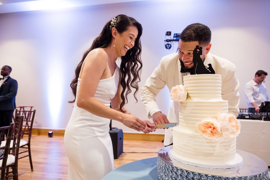 A bride and groom, smiling and laughing, cut their white wedding cake together. The cake has peach flowers and a silhouette topper. Other guests are visible in the background.