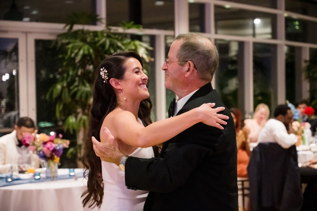 A bride in a white dress dances with an older man in a suit, both smiling happily at each other in a warmly lit reception hall with guests seated at decorated tables in the background.