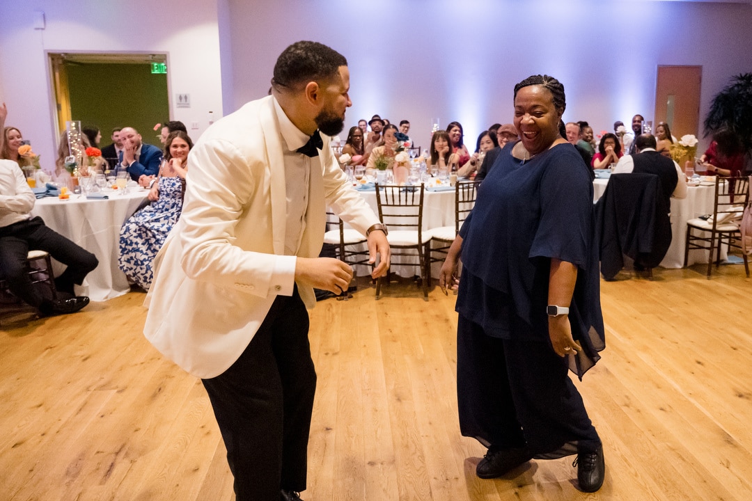Two people, one in a white tuxedo and one in a navy outfit, joyfully dance together on a wooden floor at a festive event, with smiling guests seated at round tables in the background.