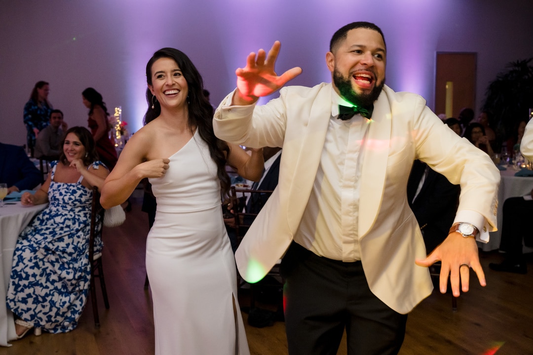 A woman in a white dress and a man in a white jacket smile and dance energetically at a party, with other guests seated and colorful lights in the background.