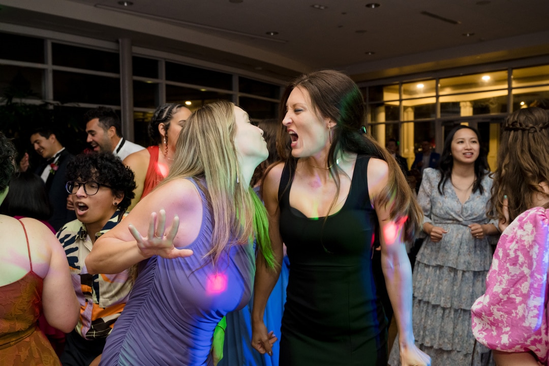 Two women in colorful dresses laugh and sing loudly while dancing at a lively indoor party. Other guests in festive attire dance and mingle in the background under bright, colorful lights.