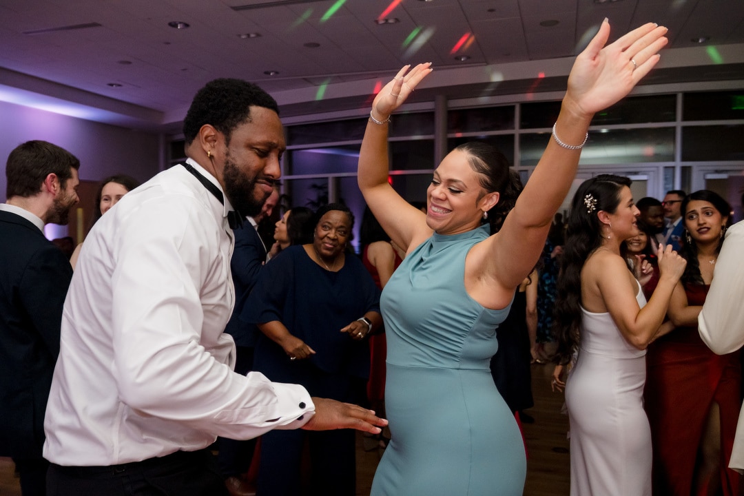 A man in a white shirt and a woman in a teal dress happily dance with arms raised at a lively indoor event, surrounded by smiling people dressed in formal attire.