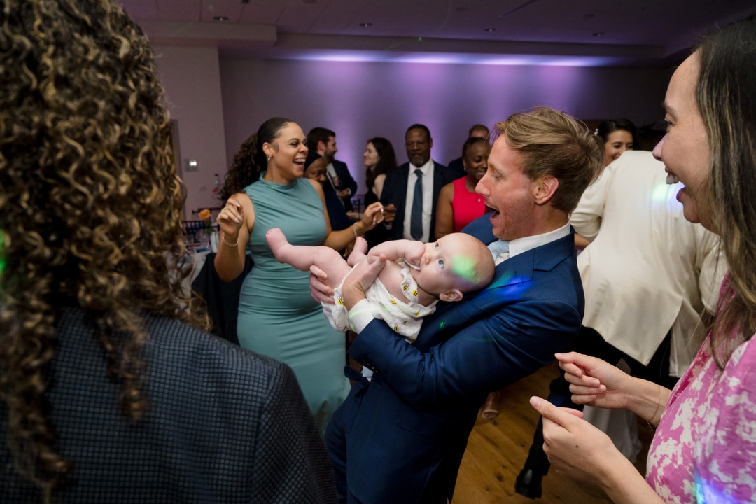 A group of adults joyfully dancing at a party, with a man in a blue suit holding a smiling baby in the center of the scene. Colorful lights and a lively atmosphere fill the room.