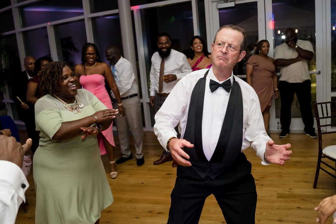 A man in a tuxedo enthusiastically dances at a lively indoor party while a group of smiling guests in colorful attire watch and join in on the wooden dance floor.