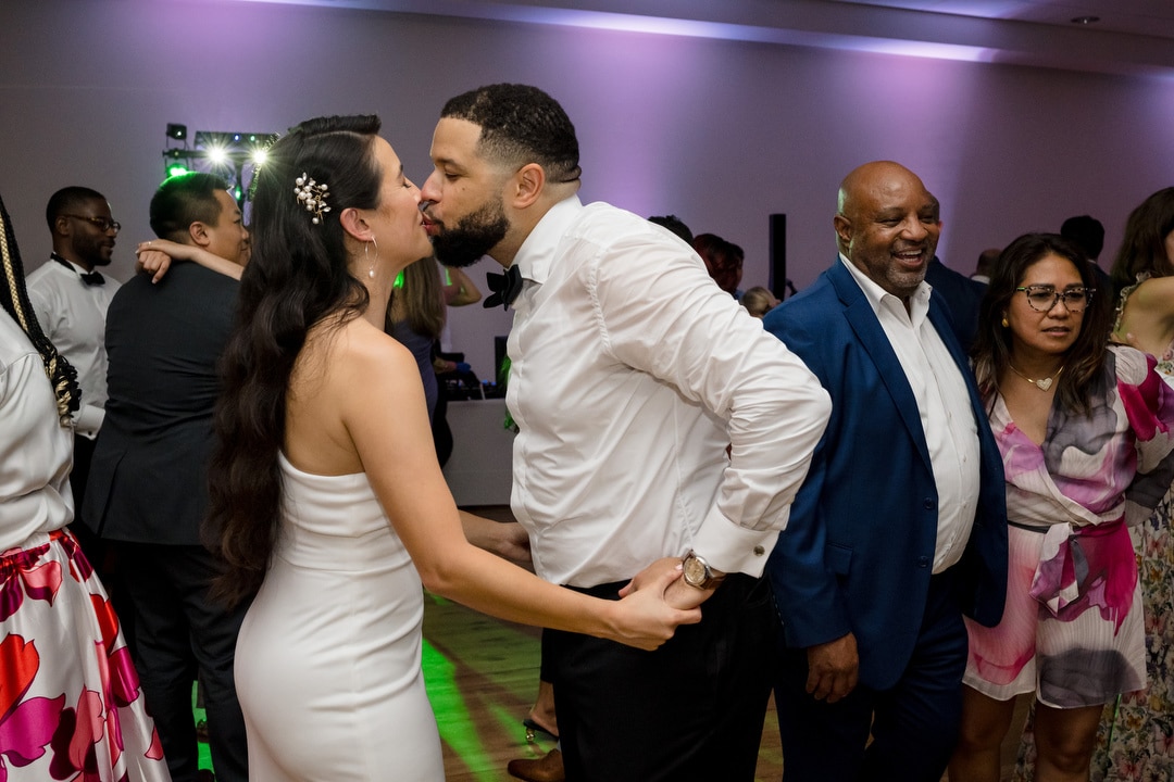 A bride and groom share a close moment on the dance floor, holding hands and smiling. Guests in colorful outfits dance and mingle in the background under purple lighting at a lively celebration.