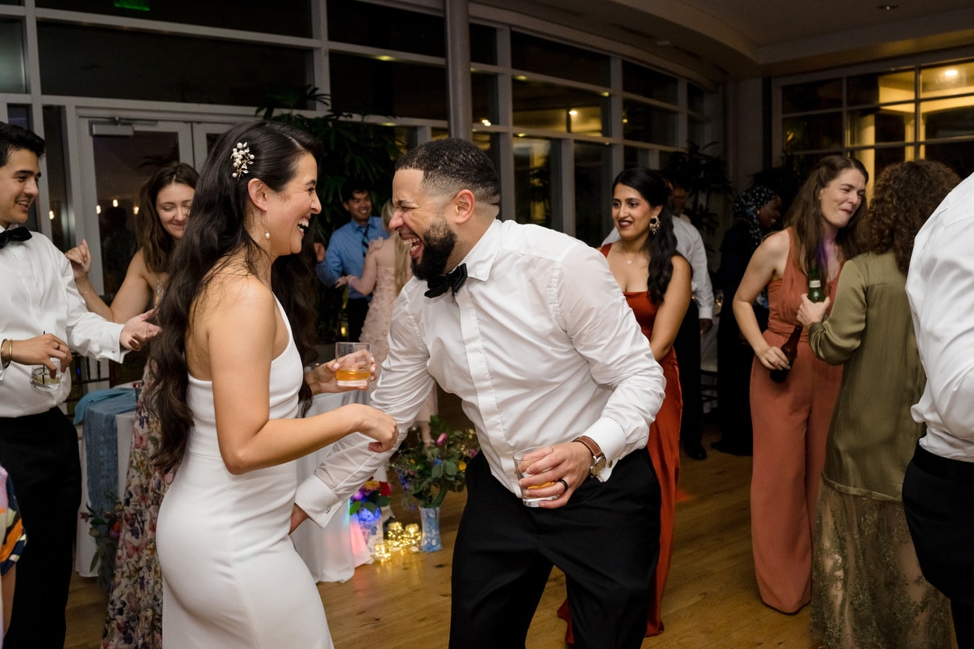 A bride in a white dress and a man in a white shirt and bow tie laugh and dance together at a lively indoor wedding reception with guests smiling and mingling in the background.