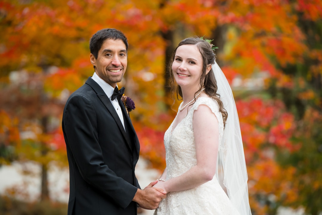 A bride and groom stand outdoors, holding hands and smiling at the camera. The bride wears a white lace dress and veil, and the groom is in a black tuxedo. Bright autumn foliage fills the background.