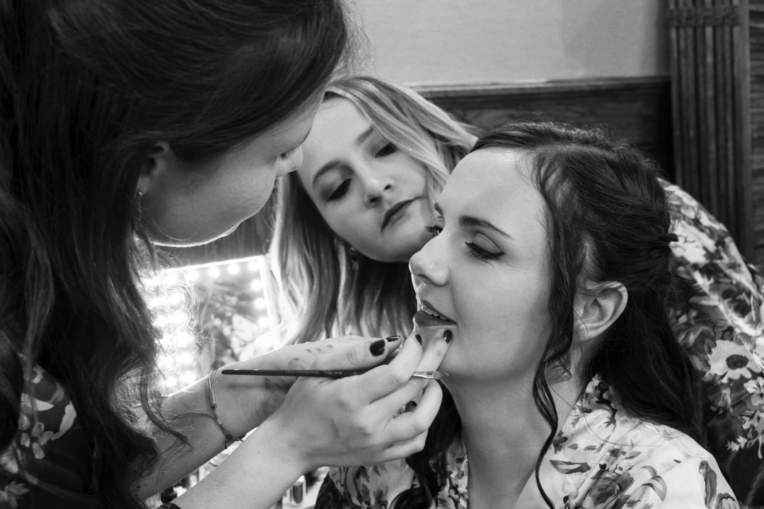 A makeup artist applies lipstick to a smiling woman while another woman looks on closely, all in a well-lit room. The atmosphere appears friendly and focused.