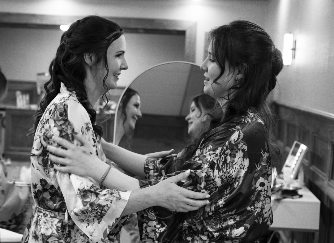 Two women in floral robes face each other, smiling and holding each other’s arms in a warmly lit room. In the background, a mirror reflects another smiling woman also wearing a robe. The setting appears joyful and intimate.