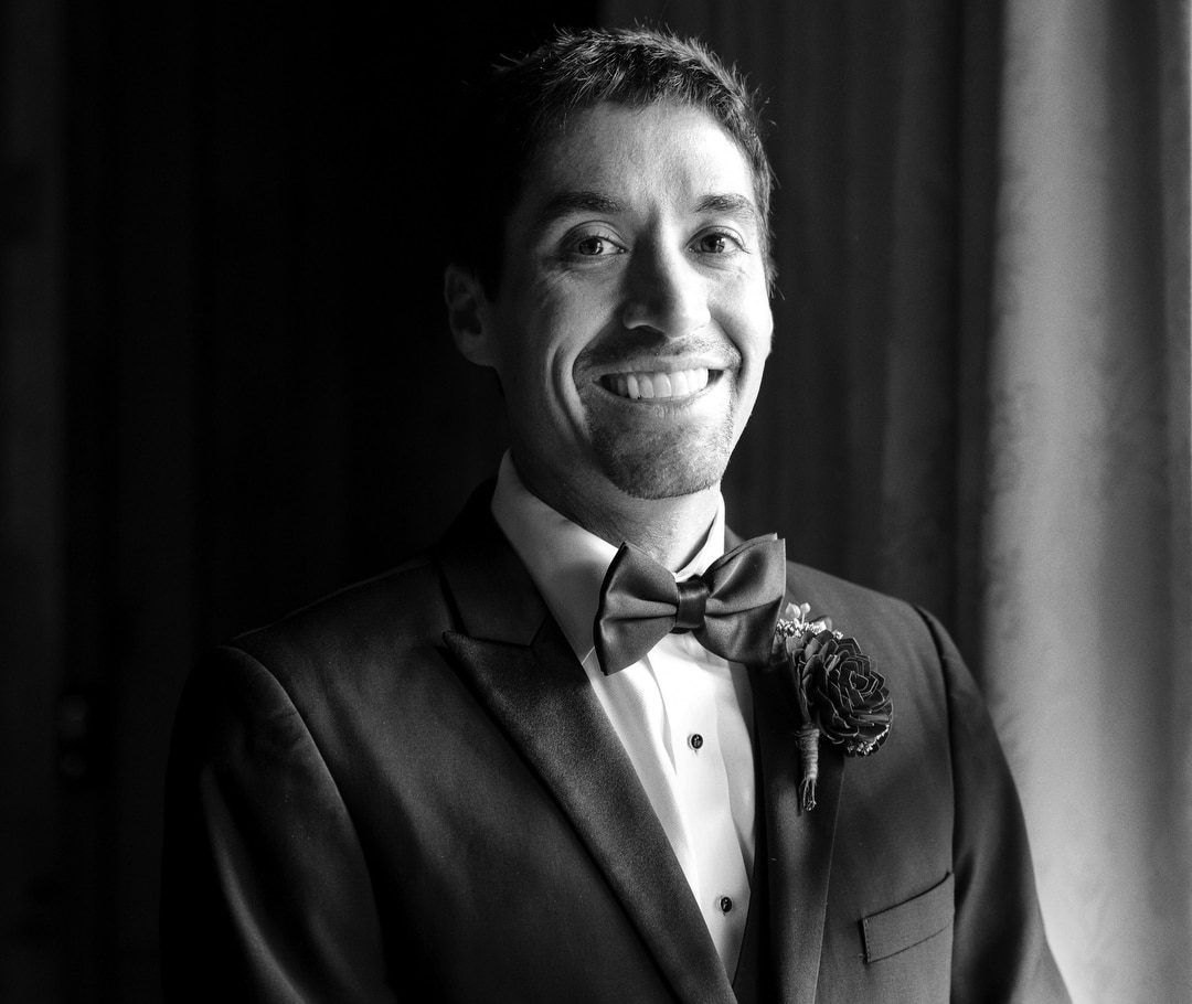 A man in a tuxedo with a bow tie smiles at the camera in black and white. He has short dark hair and a boutonniere on his lapel, standing near a softly lit curtain.
