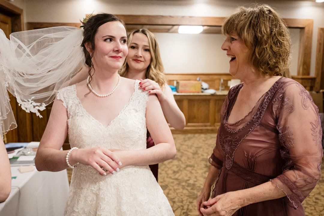 A bride in a white lace dress stands smiling as a bridesmaid adjusts her veil. A woman in a mauve dress smiles at them in a warmly lit room with wood paneling.