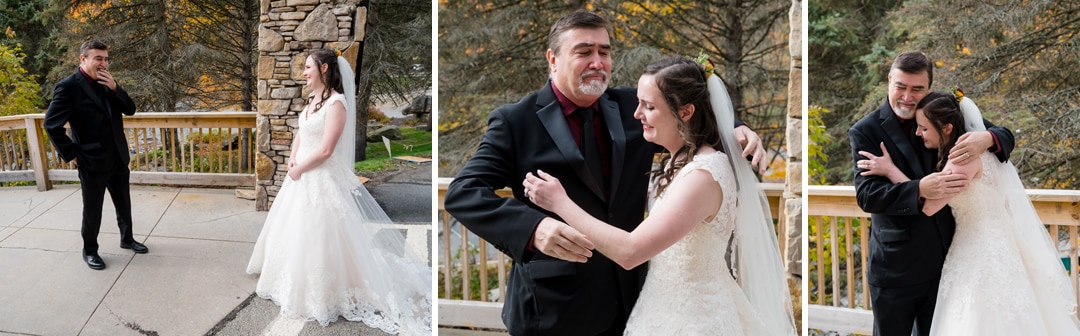 A bride in a white dress shares an emotional first look and warm hug with her father, who is wearing a black suit, outdoors by a wooden railing and stone structure.