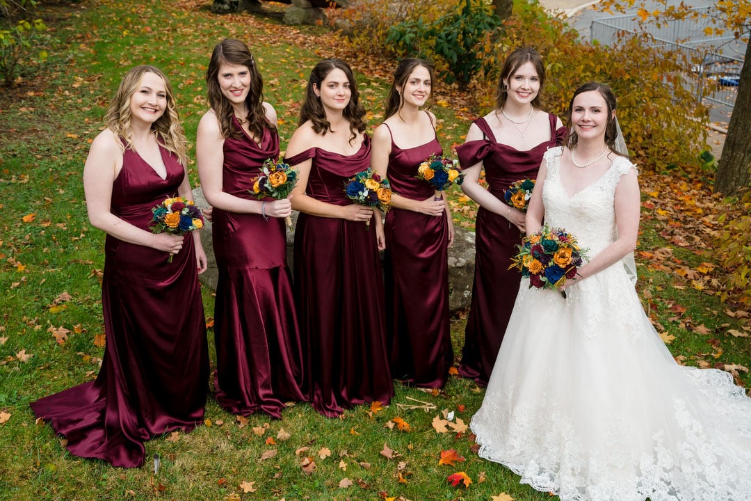 Six women stand outdoors on grass. Five bridesmaids wear matching burgundy dresses and hold bouquets, while one bride in a white gown stands at the front, also holding a bouquet. Autumn leaves are scattered on the ground.