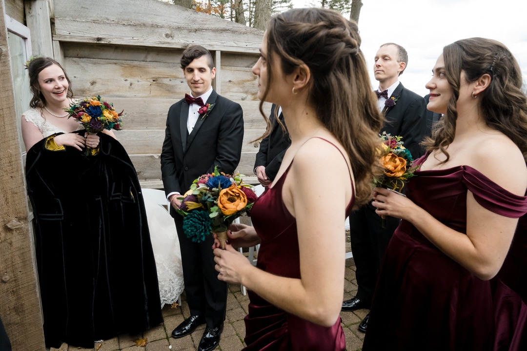 A wedding party stands outdoors. The bride in a black cloak and white dress holds a bouquet and smiles at the groom, who wears a black tuxedo. Bridesmaids in burgundy dresses and a groomsman in a tuxedo hold colorful bouquets.