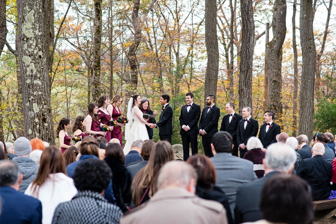 An outdoor wedding ceremony in a forested area, with a bride and groom standing together, surrounded by bridesmaids in burgundy dresses and groomsmen in black suits, as guests watch from wooden benches.