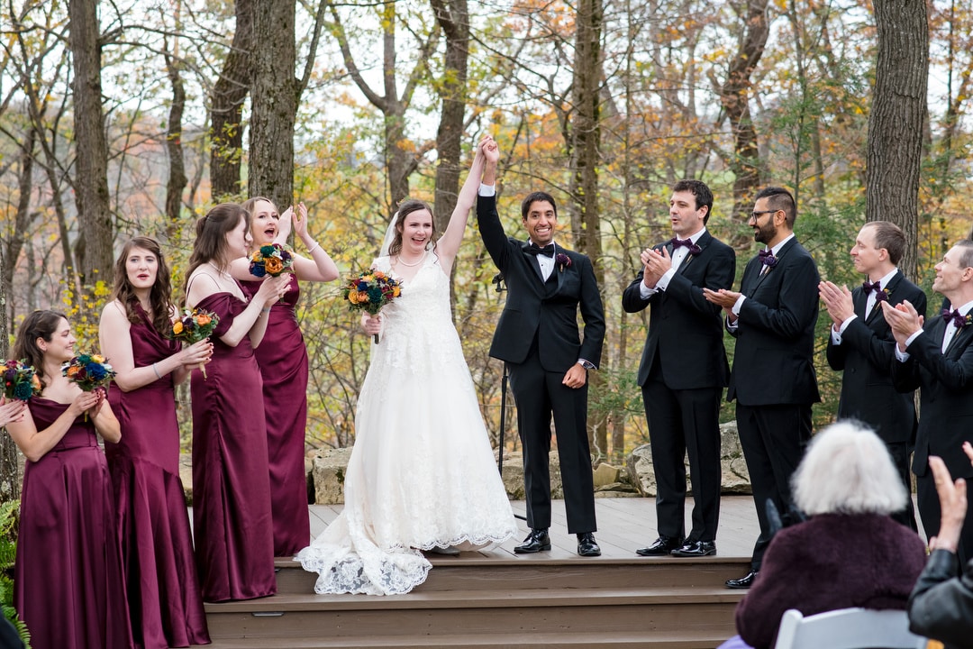 A bride in a white dress raises her arm joyfully next to the groom as bridesmaids in burgundy dresses and groomsmen in black tuxedos cheer and clap outdoors amid trees.