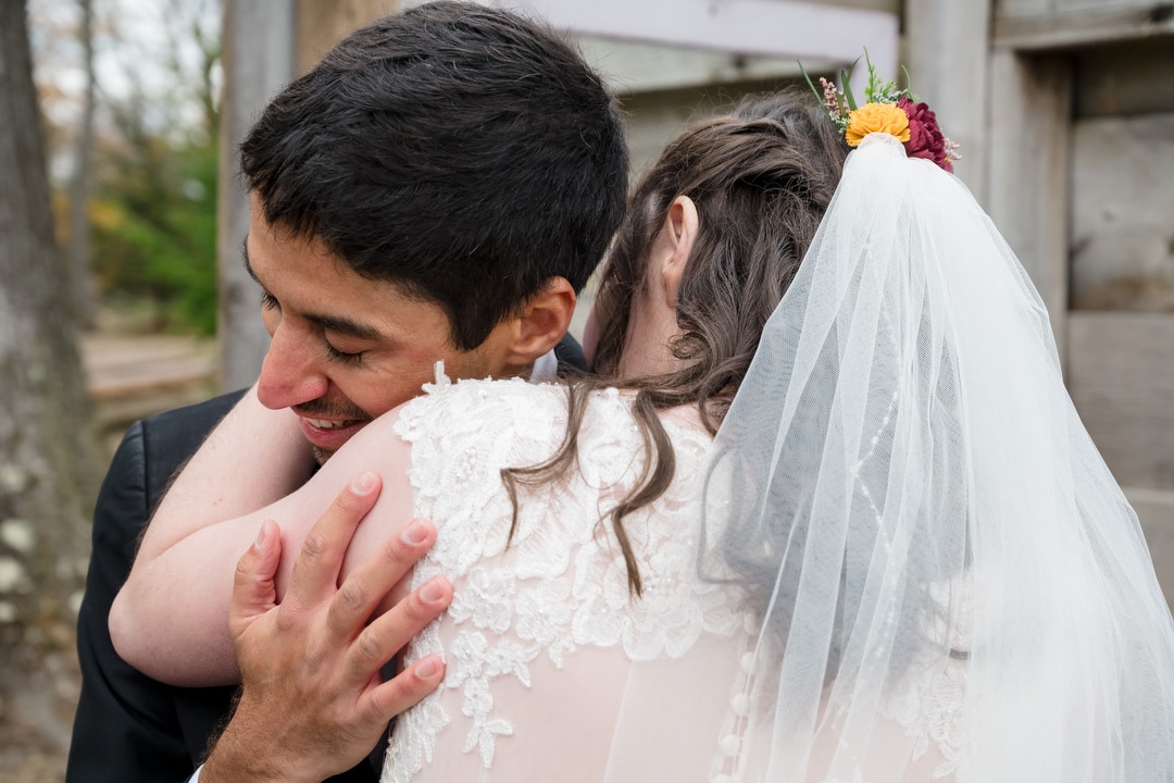 A bride and groom share a joyful, close embrace outdoors. The groom smiles with his eyes closed while hugging the bride, who wears a veil with small flowers in her hair. Their happiness is evident in the candid moment.