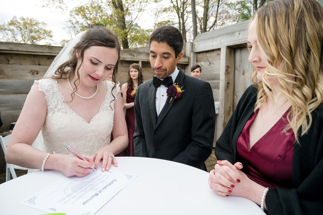A bride in a white dress signs a marriage certificate at an outdoor table, while the groom in a black tuxedo and a bridesmaid in a burgundy dress watch closely. Trees and wooden walls are in the background.