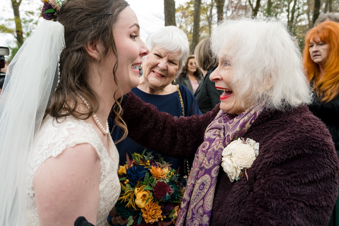 A joyful bride in a white dress and veil laughs with an elderly woman in a purple coat and scarf, surrounded by smiling guests outdoors. The elderly woman has a flower pinned to her coat and touches the brides shoulder.