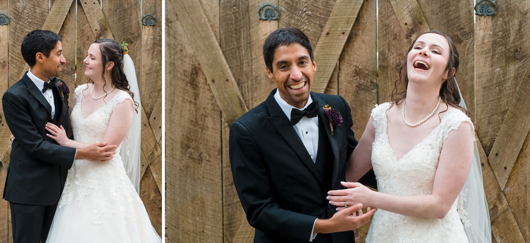 A bride in a white dress and a groom in a black tuxedo stand together smiling and laughing in front of a rustic wooden door. They are embracing and clearly enjoying a joyful moment.