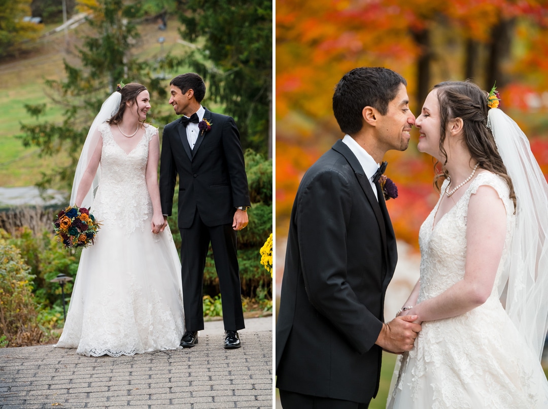 A bride in a white gown and veil holds hands and smiles with a groom in a black tuxedo. They stand outdoors, surrounded by greenery and autumn trees with vibrant orange and red leaves.
