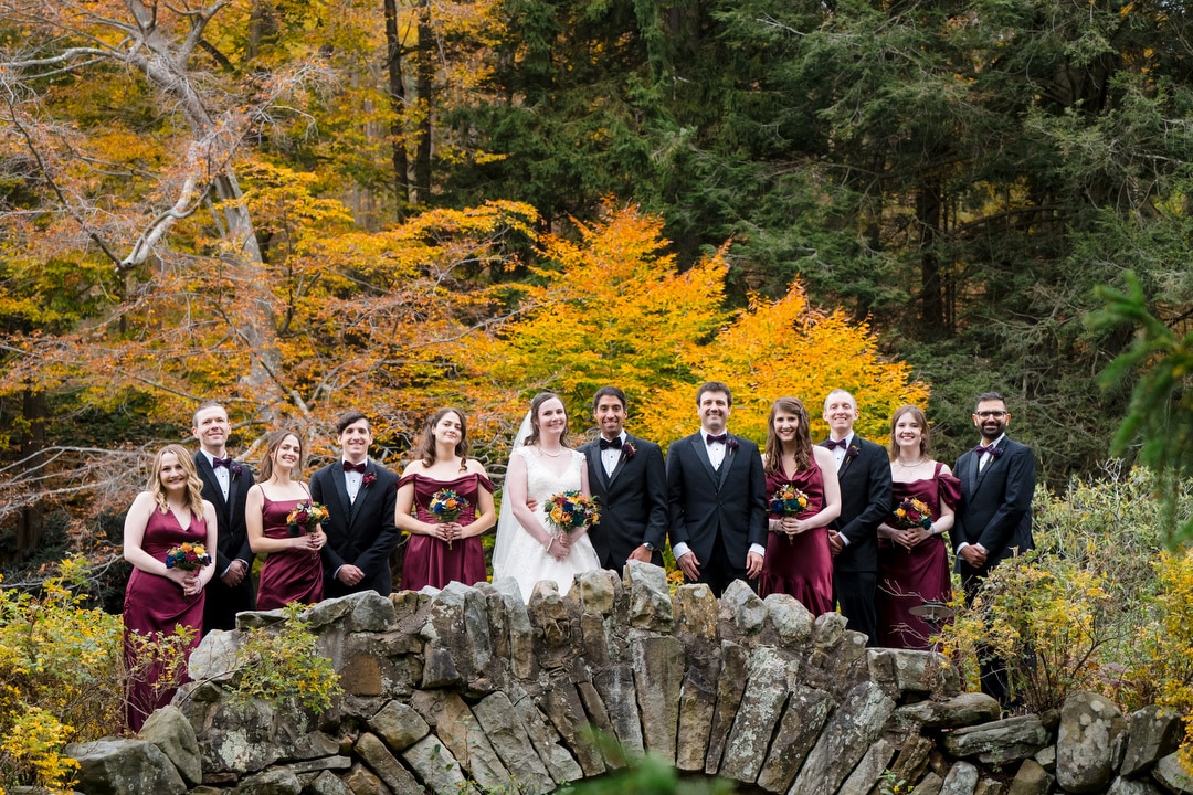 A wedding party in formal attire stands on a stone bridge surrounded by autumn foliage, with the bride and groom in the center and bridesmaids and groomsmen on either side, smiling at the camera.