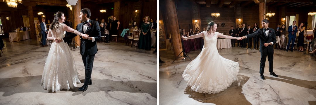 A bride and groom, dressed in formal wedding attire, dance together in a spacious, rustic venue with wooden beams and chandeliers, as guests watch and smile in the background.