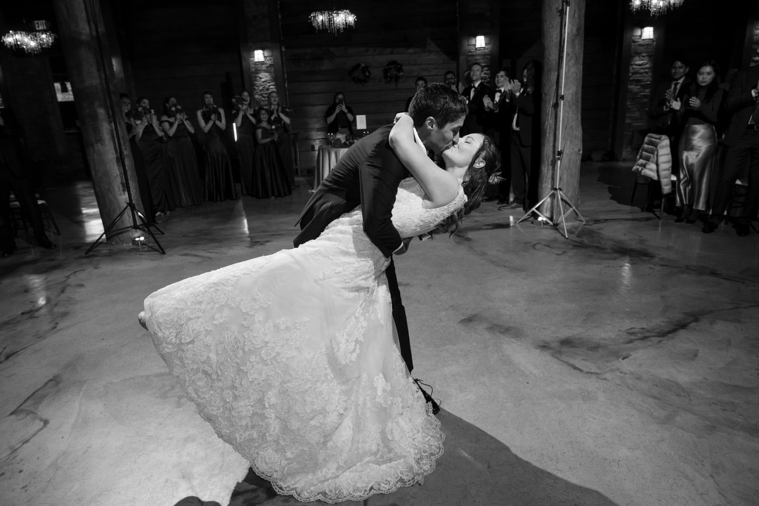 A groom dips and kisses his bride on the dance floor during their wedding, while guests in formal attire watch and smile in the background. The setting is a rustic, dimly lit venue.