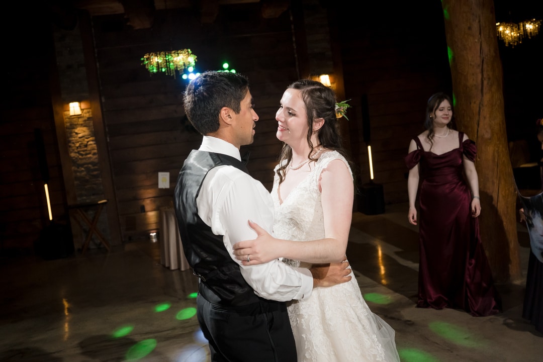 A bride and groom smile at each other while dancing together on a dimly lit, rustic wooden floor. A woman in a burgundy dress stands in the background, watching them.