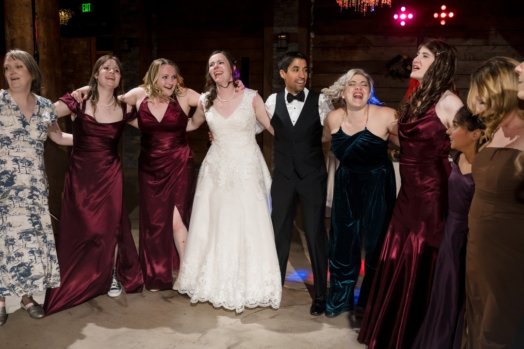 A bride and groom stand arm-in-arm with friends in formal dresses, smiling and enjoying themselves on a dance floor at a wedding reception. Colorful lights glow in the background.