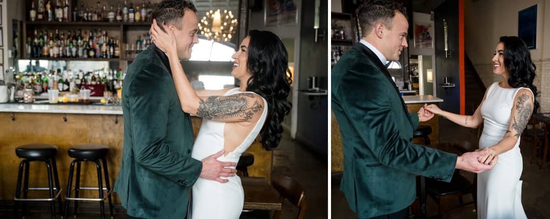A couple dressed formally, with the woman in a white gown and the man in a green velvet jacket, stand smiling and embracing each other in a stylish bar setting.
