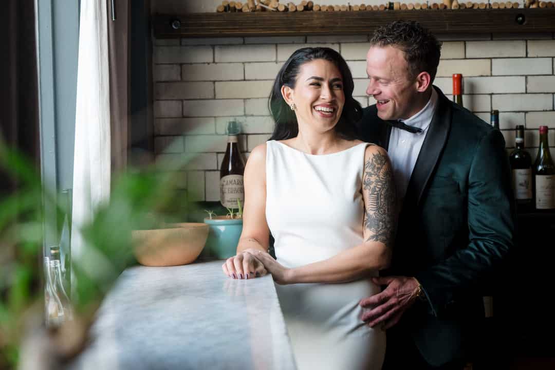 A smiling couple in formal attire stands at a marble counter in front of a tiled wall and shelves with wine bottles, sharing a joyful moment together. A plant is visible in the foreground.