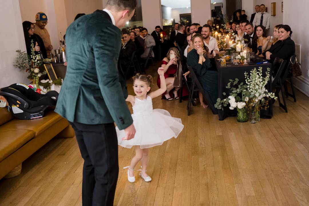 A man in a green suit jacket dances with a smiling young girl in a white dress in front of seated guests at a warmly lit indoor event, likely a wedding or party. The guests watch and smile, surrounded by candles and flowers.
