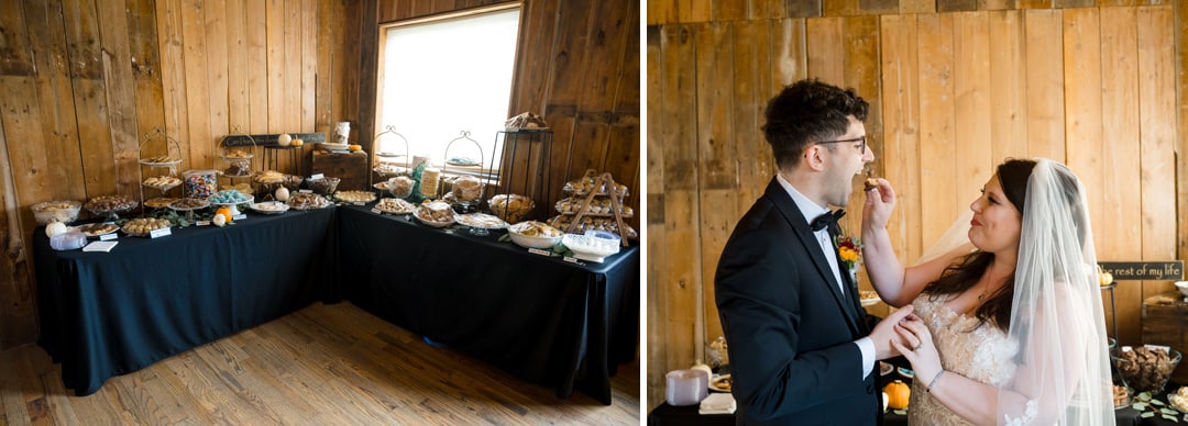 A dessert table with various treats is set up against a wooden wall. Next to it, a groom in a suit feeds a piece of dessert to his bride in a wedding dress. Both appear happy and smiling.